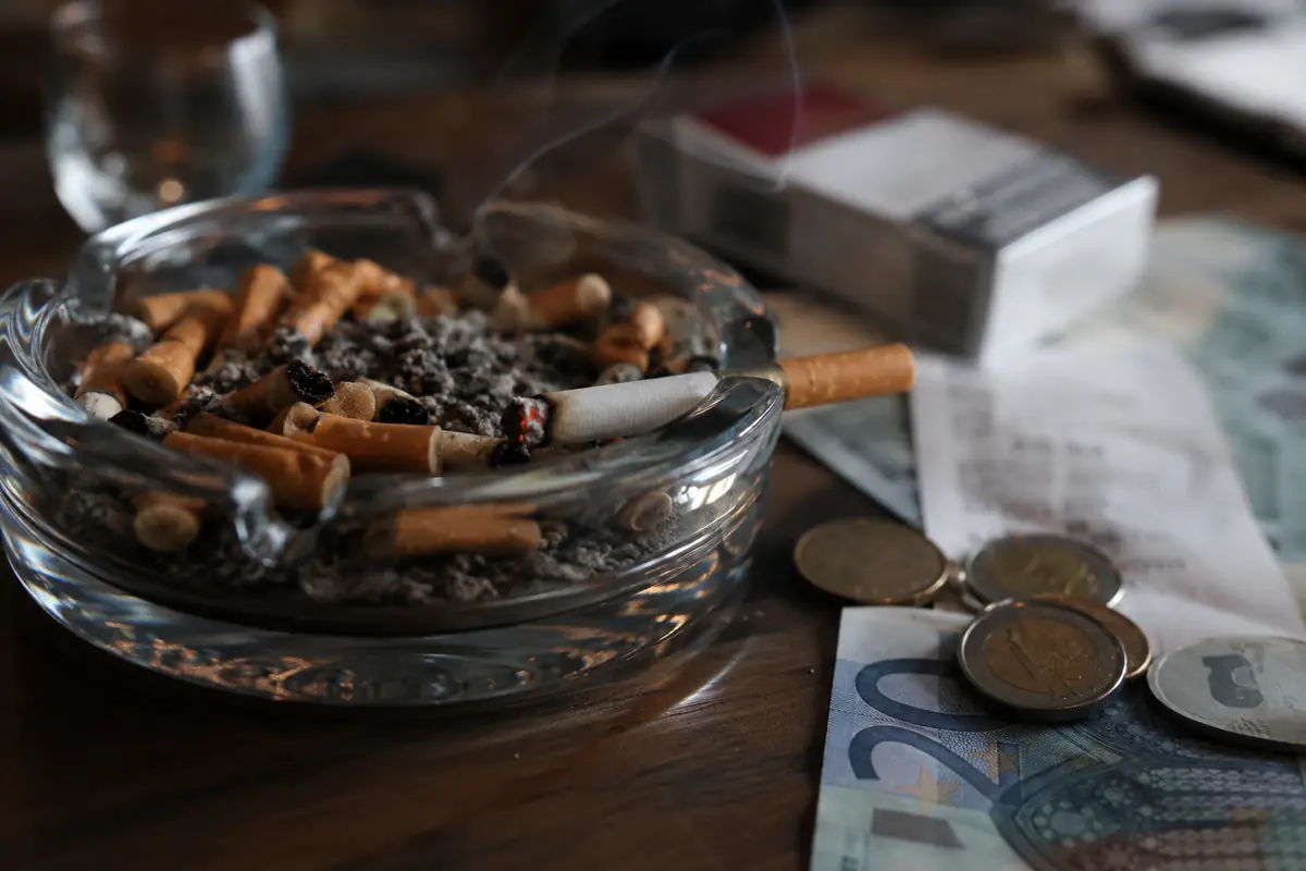 Posacenere con cicche di sigarette, una sigaretta accesa e denaro sul tavolo per rappresentare l’aumento del prezzo del tabacco