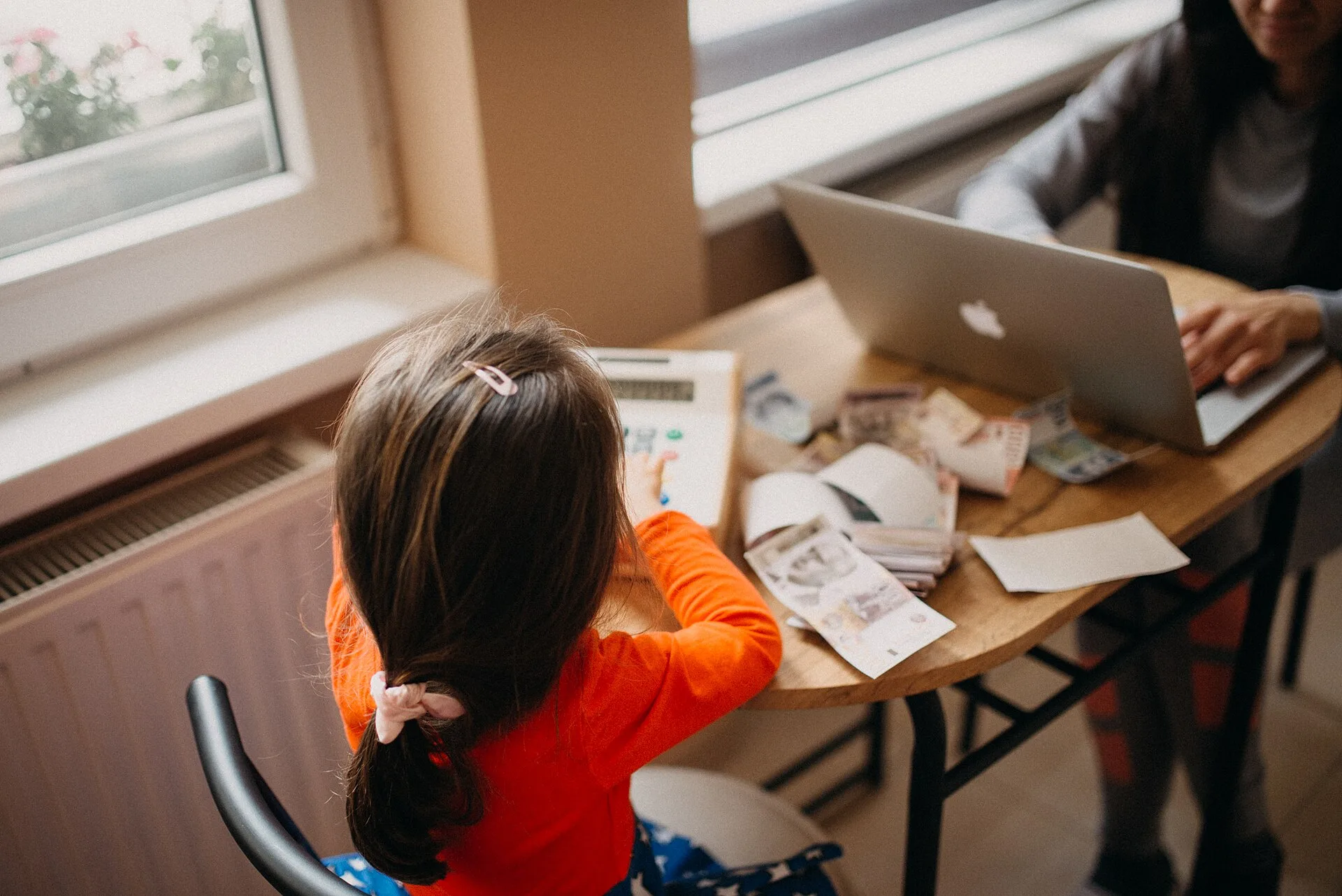 Una bambina disegna al tavolo mentre un genitore lavora al computer in casa
