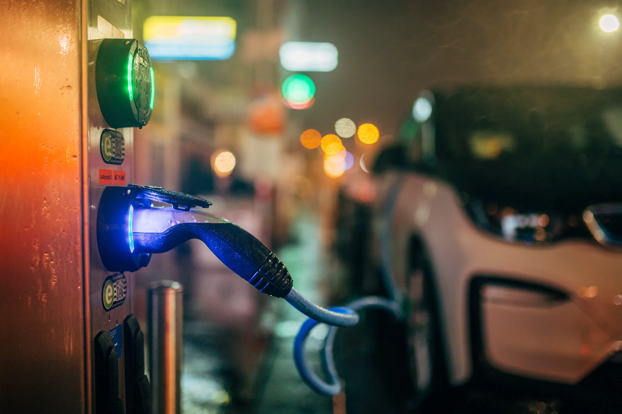 Cavo inserito in una colonnina di ricarica, con un’auto elettrica sullo sfondo